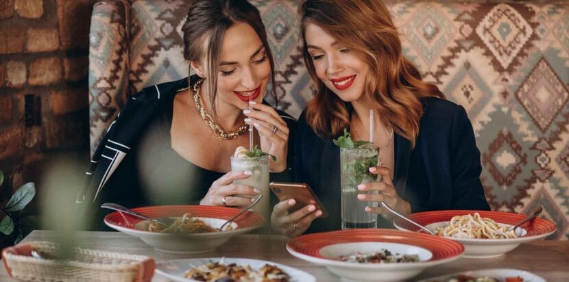 Top Indian Street Food in Alhambra, California 10 Two women sitting in a cozy restaurant booth, enjoying drinks and looking at a smartphone together, smiling and engaged. Various plates of pasta and appetizers are on the table.